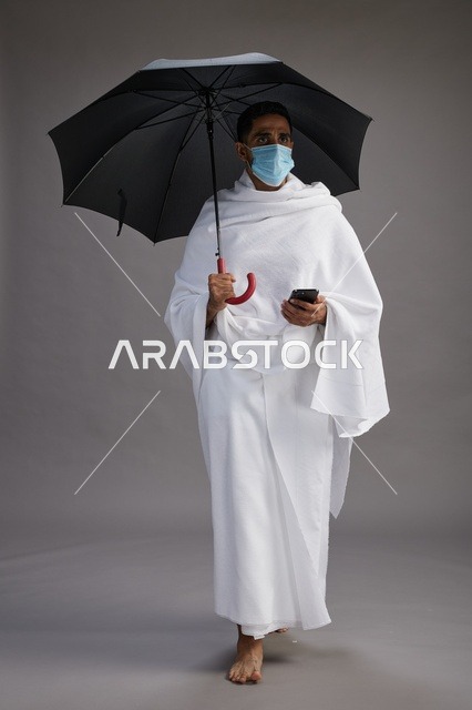 A Saudi Arabian Gulf pilgrim in Ihram clothing holding an umbrella and wearing a mask, using a mobile phone, performing the rituals of Hajj and Umrah, full body, gray background.