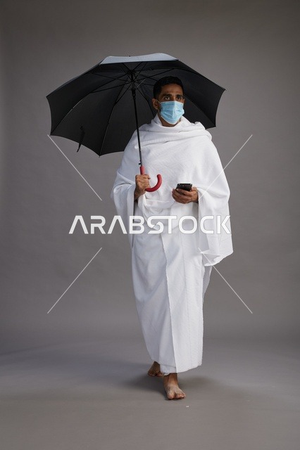 A Saudi Arabian Gulf pilgrim in Ihram clothing holding an umbrella and wearing a mask, using a mobile phone, performing the rituals of Hajj and Umrah, full body, gray background.