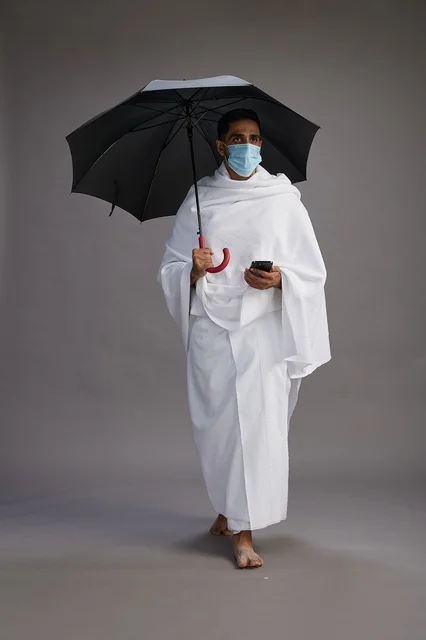 حاج  عربي سعودي خليجي بملابس الإحرام يحمل مظلة ويرتدي كمامة, استخدام الهاتف المحمول , اداء مناسك الحج والعمره, كامل الجسم, خلفيه رماديه