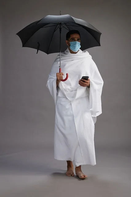 A Saudi Arabian Gulf pilgrim in Ihram clothing holding an umbrella and wearing a mask, using a mobile phone, performing the rituals of Hajj and Umrah, full body, gray background.