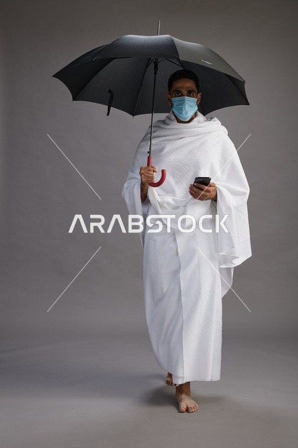 حاج  عربي سعودي خليجي بملابس الإحرام يحمل مظلة ويرتدي كمامة, استخدام الهاتف المحمول , اداء مناسك الحج والعمره, كامل الجسم, خلفيه رماديه