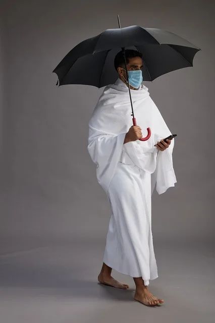 حاج  عربي سعودي خليجي بملابس الإحرام يحمل مظلة ويرتدي كمامة, استخدام الهاتف المحمول , اداء مناسك الحج والعمره, كامل الجسم, خلفيه رماديه