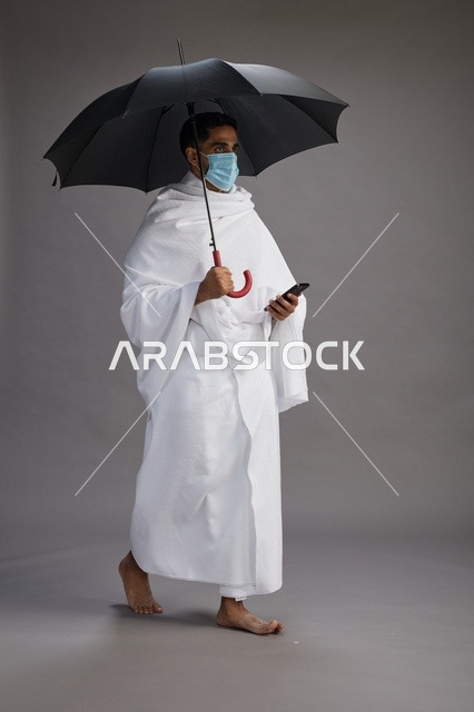 A Saudi Arabian Gulf pilgrim in Ihram clothing holding an umbrella and wearing a mask, using a mobile phone, performing the rituals of Hajj and Umrah, full body, gray background.