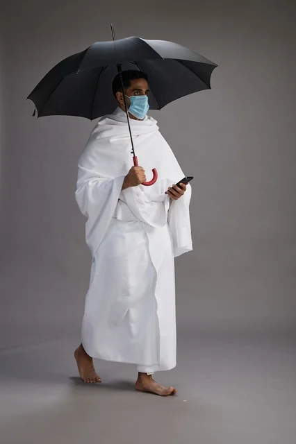 A Saudi Arabian Gulf pilgrim in Ihram clothing holding an umbrella and wearing a mask, using a mobile phone, performing the rituals of Hajj and Umrah, full body, gray background.