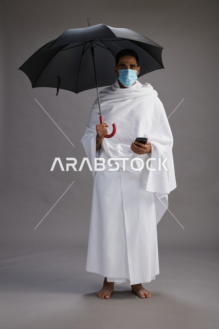 حاج  عربي سعودي خليجي بملابس الإحرام يحمل مظلة ويرتدي كمامة, استخدام الهاتف المحمول , اداء مناسك الحج والعمره, كامل الجسم, خلفيه رماديه