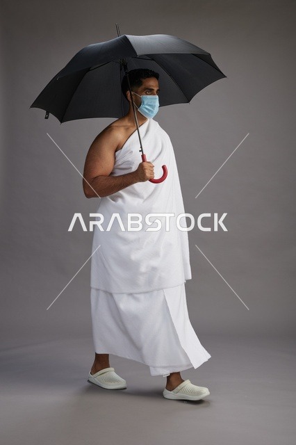 A portrait of an Arab Saudi Gulf pilgrim in Ihram clothing holding an umbrella and wearing a mask for protection against COVID-19, full body, gray background.