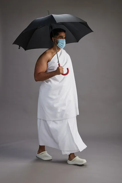 بورتريه لحاج  عربي سعودي  خليجي بملابس الإحرام يحمل مظلة ويرتدي كمامة, الوقايه من كوفيد 19, كامل الجسم, خلفيه رماديه