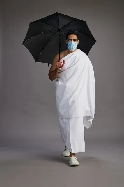 بورتريه لحاج  عربي سعودي  خليجي بملابس الإحرام يحمل مظلة ويرتدي كمامة, الوقايه من كوفيد 19, كامل الجسم, خلفيه رماديه