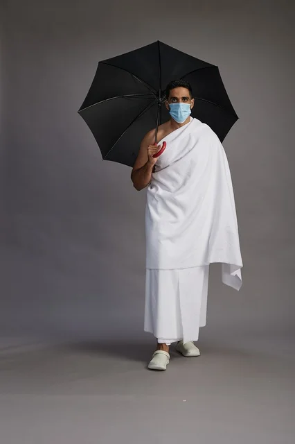 A portrait of an Arab Saudi Gulf pilgrim in Ihram clothing holding an umbrella and wearing a mask for protection against COVID-19, full body, gray background.