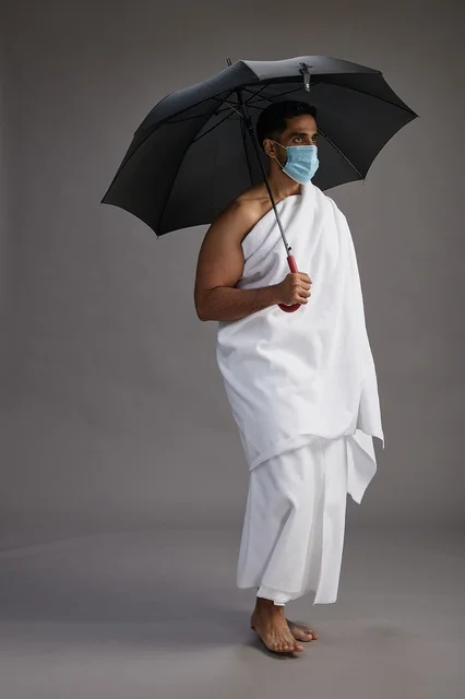 A portrait of an Arab Saudi Gulf pilgrim in Ihram clothing holding an umbrella and wearing a mask for protection against COVID-19, full body, gray background.