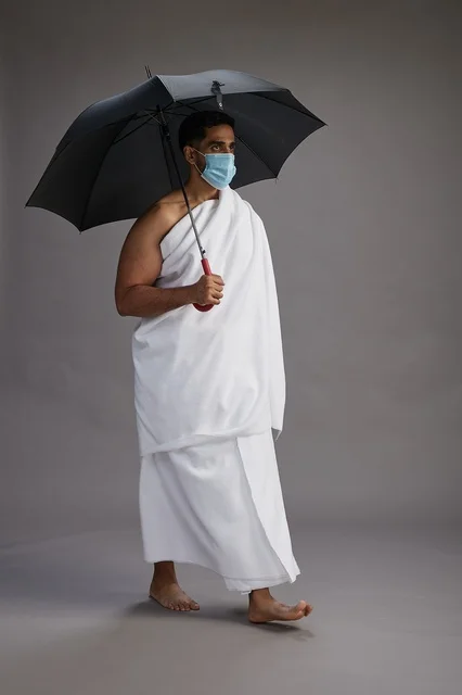 A portrait of an Arab Saudi Gulf pilgrim in Ihram clothing holding an umbrella and wearing a mask for protection against COVID-19, full body, gray background.