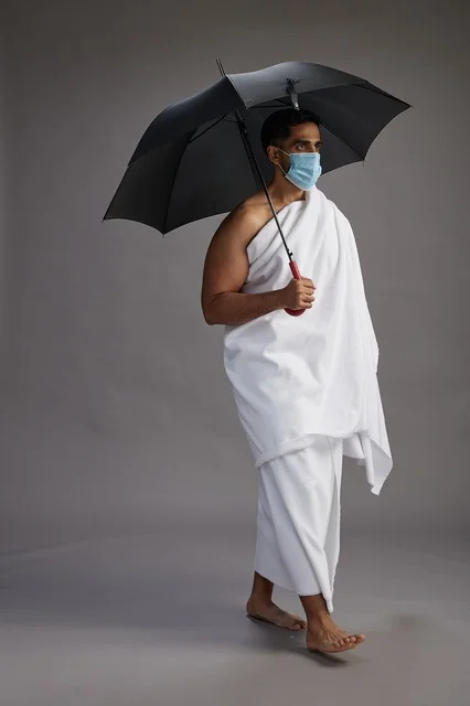 A portrait of an Arab Saudi Gulf pilgrim in Ihram clothing holding an umbrella and wearing a mask for protection against COVID-19, full body, gray background.