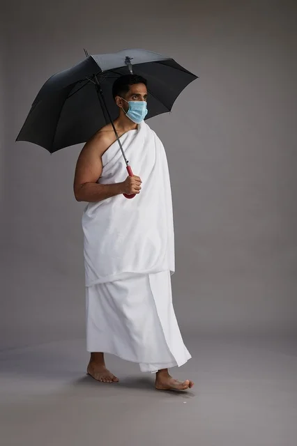بورتريه لحاج  عربي سعودي  خليجي بملابس الإحرام يحمل مظلة ويرتدي كمامة, الوقايه من كوفيد 19, كامل الجسم, خلفيه رماديه
