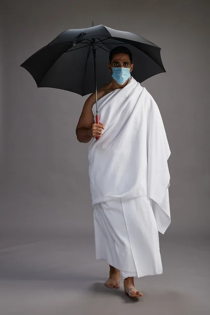 A portrait of an Arab Saudi Gulf pilgrim in Ihram clothing holding an umbrella and wearing a mask for protection against COVID-19, full body, gray background.