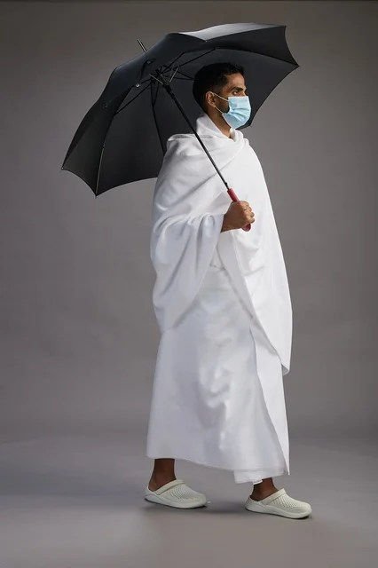 A portrait of an Arab Saudi Gulf pilgrim in Ihram clothing holding an umbrella and wearing a mask for protection against COVID-19, full body, gray background.