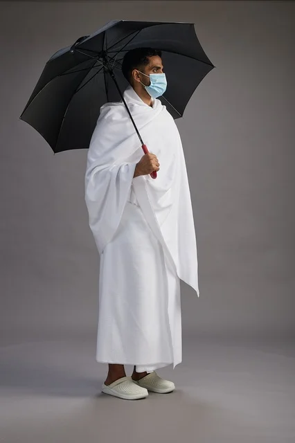 بورتريه لحاج  عربي سعودي  خليجي بملابس الإحرام يحمل مظلة ويرتدي كمامة, الوقايه من كوفيد 19, كامل الجسم, خلفيه رماديه