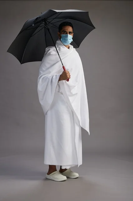 بورتريه لحاج  عربي سعودي  خليجي بملابس الإحرام يحمل مظلة ويرتدي كمامة, الوقايه من كوفيد 19, كامل الجسم, خلفيه رماديه