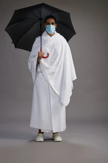 A portrait of an Arab Saudi Gulf pilgrim in Ihram clothing holding an umbrella and wearing a mask for protection against COVID-19, full body, gray background.