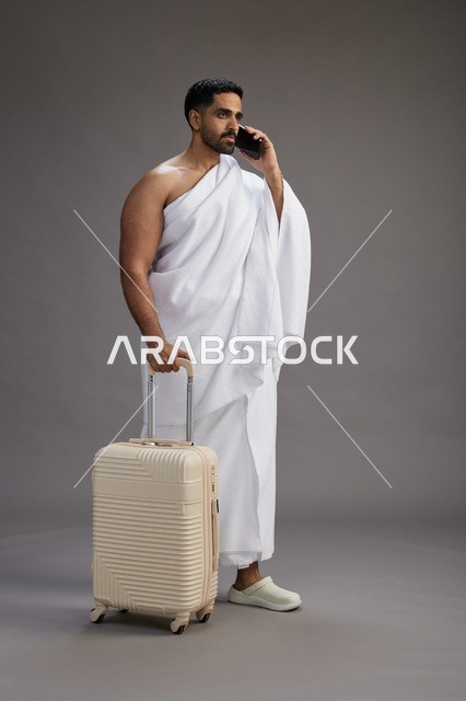 بورتريه لرجل عربي  سعودي خليجي بملابس الإحرام مع حقيبة سفر, استخدام الهاتف المحمول, الذهاب الى مكه للحج والعمره, كامل الجسم, خلفيه رماديه