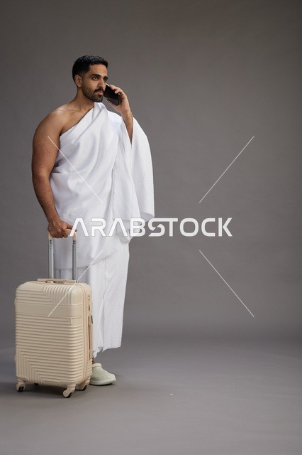 بورتريه لرجل عربي  سعودي خليجي بملابس الإحرام مع حقيبة سفر, استخدام الهاتف المحمول, الذهاب الى مكه للحج والعمره, كامل الجسم, خلفيه رماديه