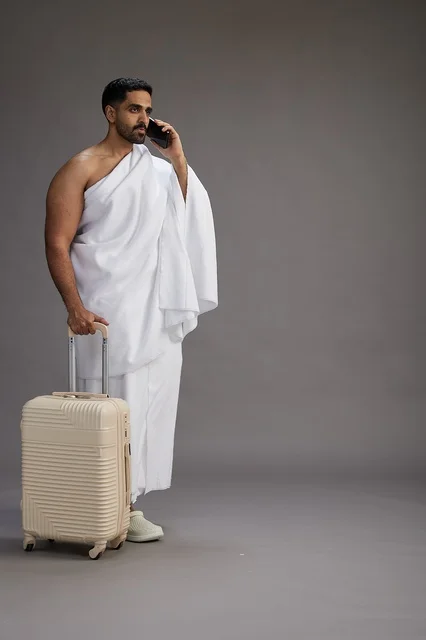 A portrait of an Arab Saudi Gulf man in Ihram clothing with a travel bag, using a mobile phone, going to Mecca for Hajj and Umrah, full body, gray background.