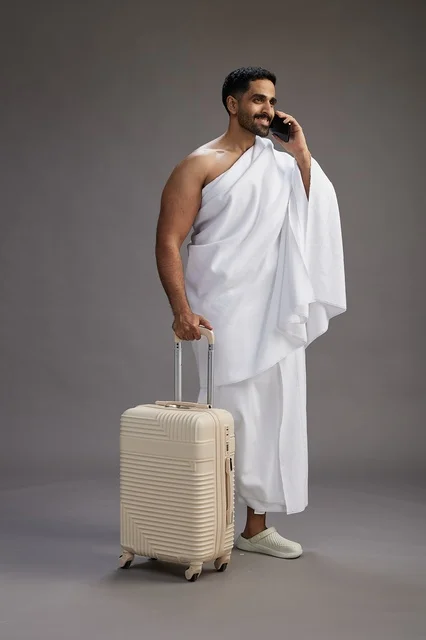 بورتريه لرجل عربي  سعودي خليجي بملابس الإحرام مع حقيبة سفر, استخدام الهاتف المحمول, الذهاب الى مكه للحج والعمره, كامل الجسم, خلفيه رماديه