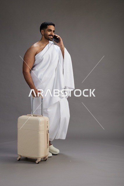 A portrait of an Arab Saudi Gulf man in Ihram clothing with a travel bag, using a mobile phone, going to Mecca for Hajj and Umrah, full body, gray background.
