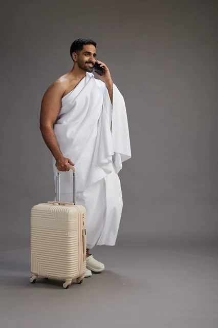 A portrait of an Arab Saudi Gulf man in Ihram clothing with a travel bag, using a mobile phone, going to Mecca for Hajj and Umrah, full body, gray background.