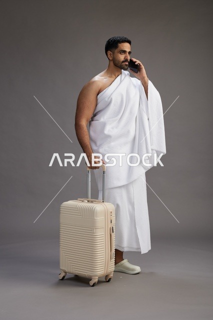 A portrait of an Arab Saudi Gulf man in Ihram clothing with a travel bag, using a mobile phone, going to Mecca for Hajj and Umrah, full body, gray background.