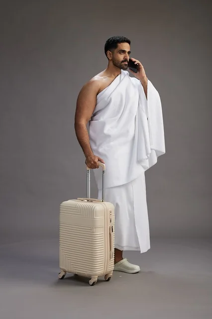بورتريه لرجل عربي  سعودي خليجي بملابس الإحرام مع حقيبة سفر, استخدام الهاتف المحمول, الذهاب الى مكه للحج والعمره, كامل الجسم, خلفيه رماديه