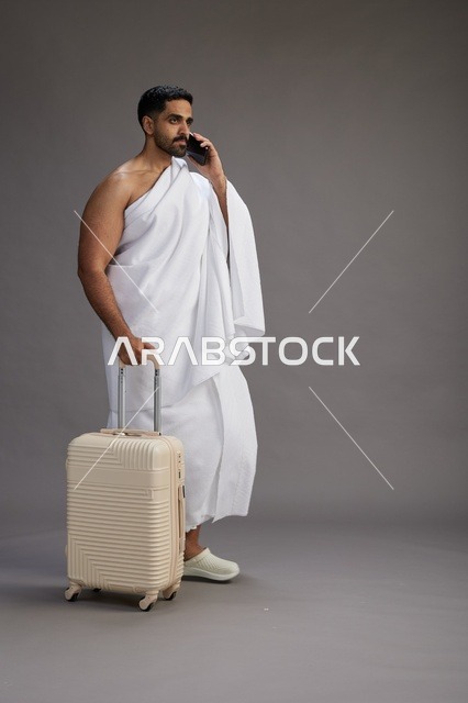 بورتريه لرجل عربي  سعودي خليجي بملابس الإحرام مع حقيبة سفر, استخدام الهاتف المحمول, الذهاب الى مكه للحج والعمره, كامل الجسم, خلفيه رماديه