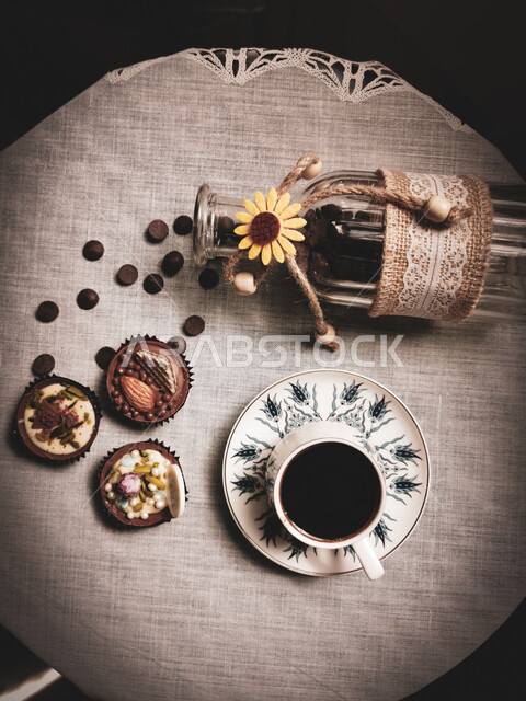 صورة من أعلى لفنجان قهوة بجانب علبة بن زجاجية رائعة مع حبوب المتناثرة على سطح الطاولة مع مجموعة من الحلويات و السكاكر اللذيذة ، خلفية سوداء 