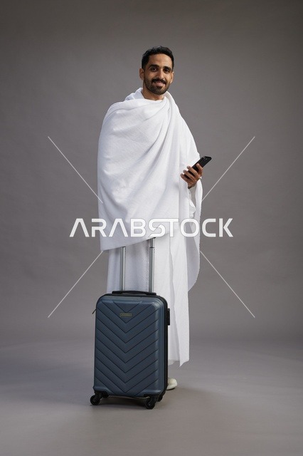 بورتريه لرجل عربي  سعودي خليجي بملابس الإحرام مع حقيبة سفر, استخدام الهاتف المحمول, الذهاب الى مكه للحج والعمره, كامل الجسم, خلفيه رماديه