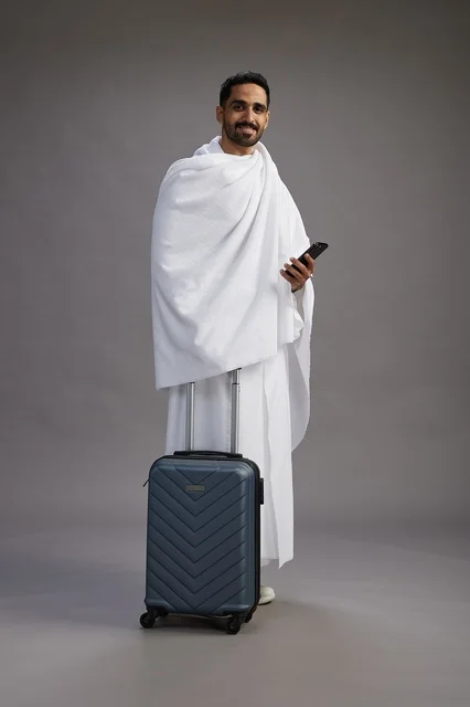 A portrait of an Arab Saudi Gulf man in Ihram clothing with a travel bag, using a mobile phone, going to Mecca for Hajj and Umrah, full body, gray background.