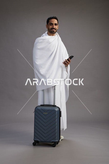 بورتريه لرجل عربي  سعودي خليجي بملابس الإحرام مع حقيبة سفر, استخدام الهاتف المحمول, الذهاب الى مكه للحج والعمره, كامل الجسم, خلفيه رماديه