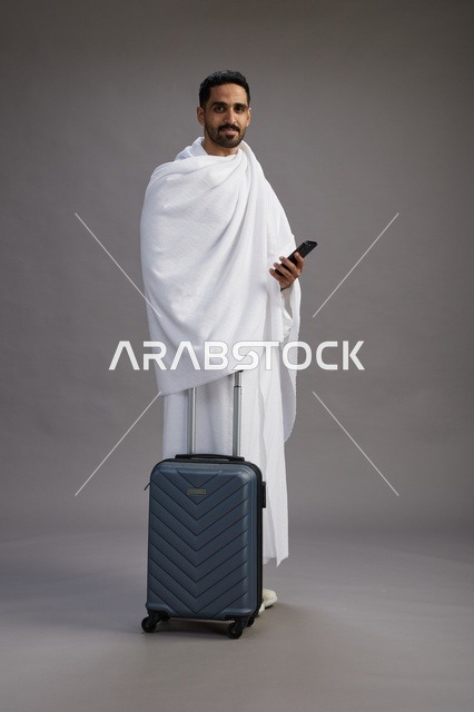 بورتريه لرجل عربي  سعودي خليجي بملابس الإحرام مع حقيبة سفر, استخدام الهاتف المحمول, الذهاب الى مكه للحج والعمره, كامل الجسم, خلفيه رماديه