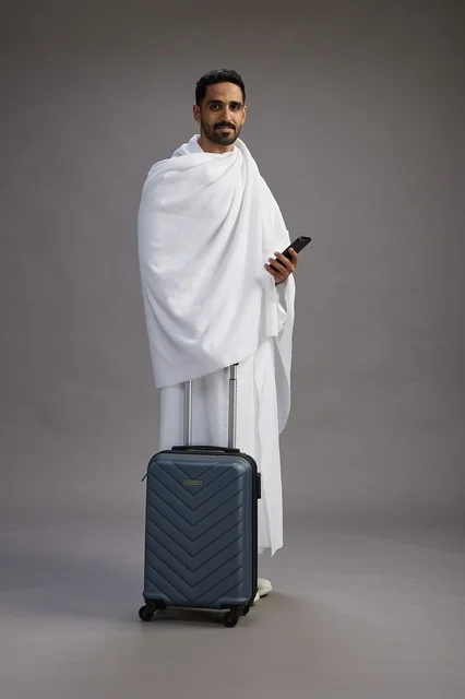 بورتريه لرجل عربي  سعودي خليجي بملابس الإحرام مع حقيبة سفر, استخدام الهاتف المحمول, الذهاب الى مكه للحج والعمره, كامل الجسم, خلفيه رماديه