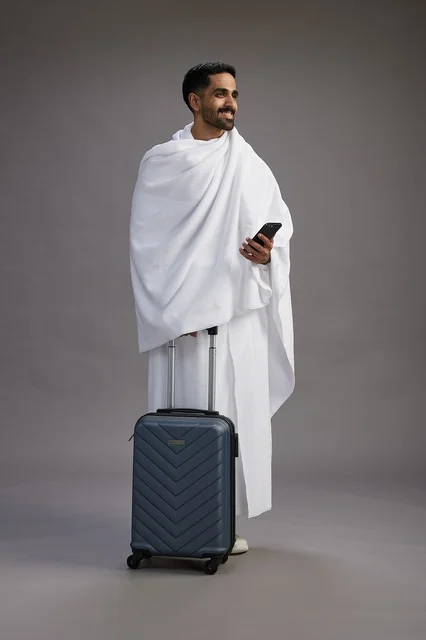 بورتريه لرجل عربي  سعودي خليجي بملابس الإحرام مع حقيبة سفر, استخدام الهاتف المحمول, الذهاب الى مكه للحج والعمره, كامل الجسم, خلفيه رماديه