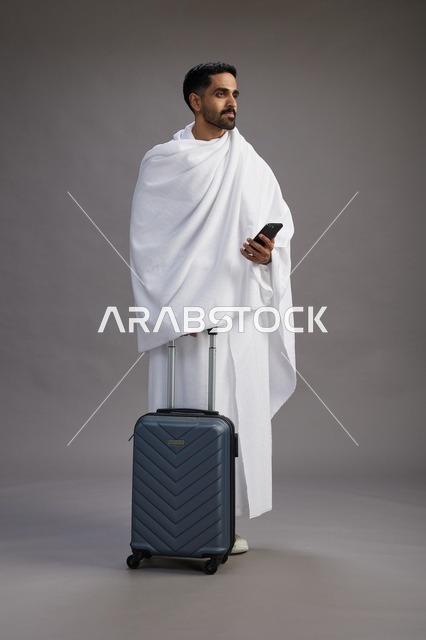 بورتريه لرجل عربي  سعودي خليجي بملابس الإحرام مع حقيبة سفر, استخدام الهاتف المحمول, الذهاب الى مكه للحج والعمره, كامل الجسم, خلفيه رماديه