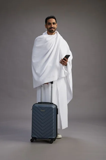 A portrait of an Arab Saudi Gulf man in Ihram clothing with a travel bag, using a mobile phone, going to Mecca for Hajj and Umrah, full body, gray background.