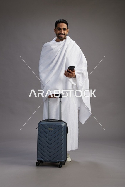 بورتريه لرجل عربي  سعودي خليجي بملابس الإحرام مع حقيبة سفر, استخدام الهاتف المحمول, الذهاب الى مكه للحج والعمره, كامل الجسم, خلفيه رماديه