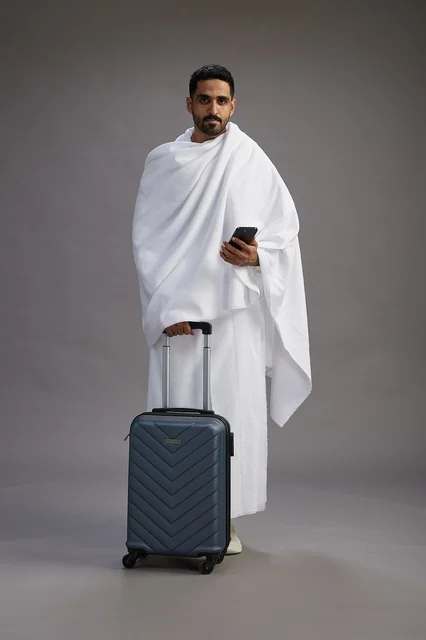 A portrait of an Arab Saudi Gulf man in Ihram clothing with a travel bag, using a mobile phone, going to Mecca for Hajj and Umrah, full body, gray background.