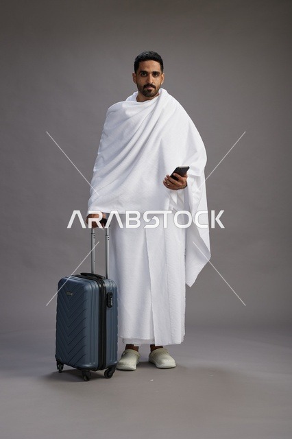 بورتريه لرجل عربي  سعودي خليجي بملابس الإحرام مع حقيبة سفر, استخدام الهاتف المحمول, الذهاب الى مكه للحج والعمره, كامل الجسم, خلفيه رماديه