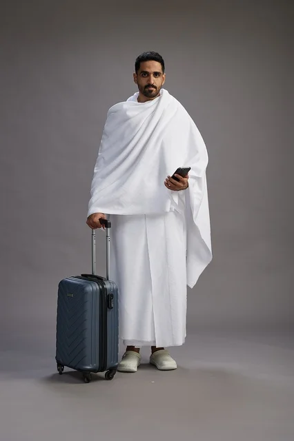 A portrait of an Arab Saudi Gulf man in Ihram clothing with a travel bag, using a mobile phone, going to Mecca for Hajj and Umrah, full body, gray background.