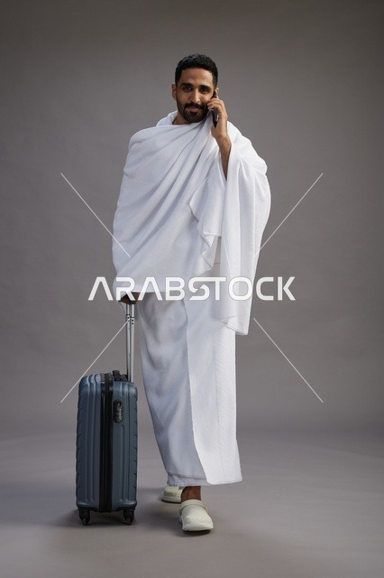 بورتريه لرجل عربي  سعودي خليجي بملابس الإحرام مع حقيبة سفر, استخدام الهاتف المحمول, الذهاب الى مكه للحج والعمره, كامل الجسم, خلفيه رماديه