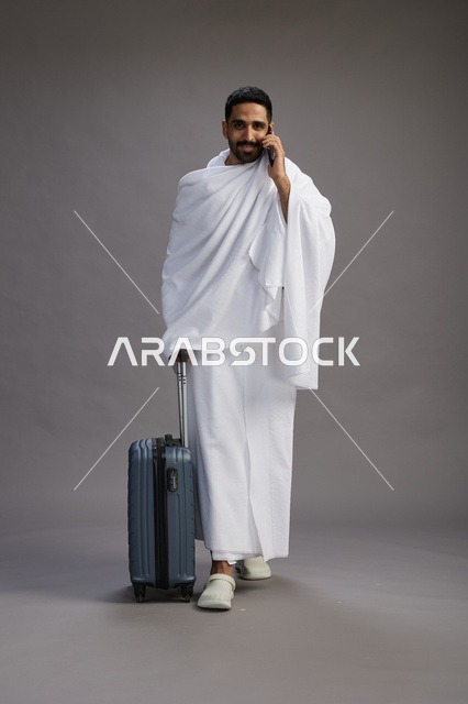 بورتريه لرجل عربي  سعودي خليجي بملابس الإحرام مع حقيبة سفر, استخدام الهاتف المحمول, الذهاب الى مكه للحج والعمره, كامل الجسم, خلفيه رماديه