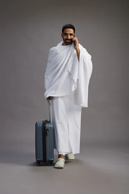 بورتريه لرجل عربي  سعودي خليجي بملابس الإحرام مع حقيبة سفر, استخدام الهاتف المحمول, الذهاب الى مكه للحج والعمره, كامل الجسم, خلفيه رماديه