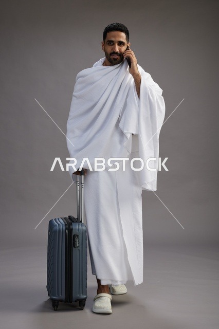 A portrait of an Arab Saudi Gulf man in Ihram clothing with a travel bag, using a mobile phone, going to Mecca for Hajj and Umrah, full body, gray background.