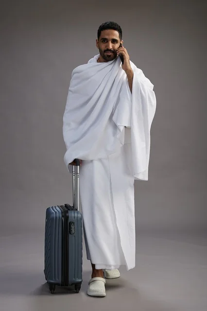 بورتريه لرجل عربي  سعودي خليجي بملابس الإحرام مع حقيبة سفر, استخدام الهاتف المحمول, الذهاب الى مكه للحج والعمره, كامل الجسم, خلفيه رماديه