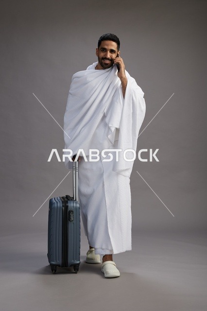 بورتريه لرجل عربي  سعودي خليجي بملابس الإحرام مع حقيبة سفر, استخدام الهاتف المحمول, الذهاب الى مكه للحج والعمره, كامل الجسم, خلفيه رماديه